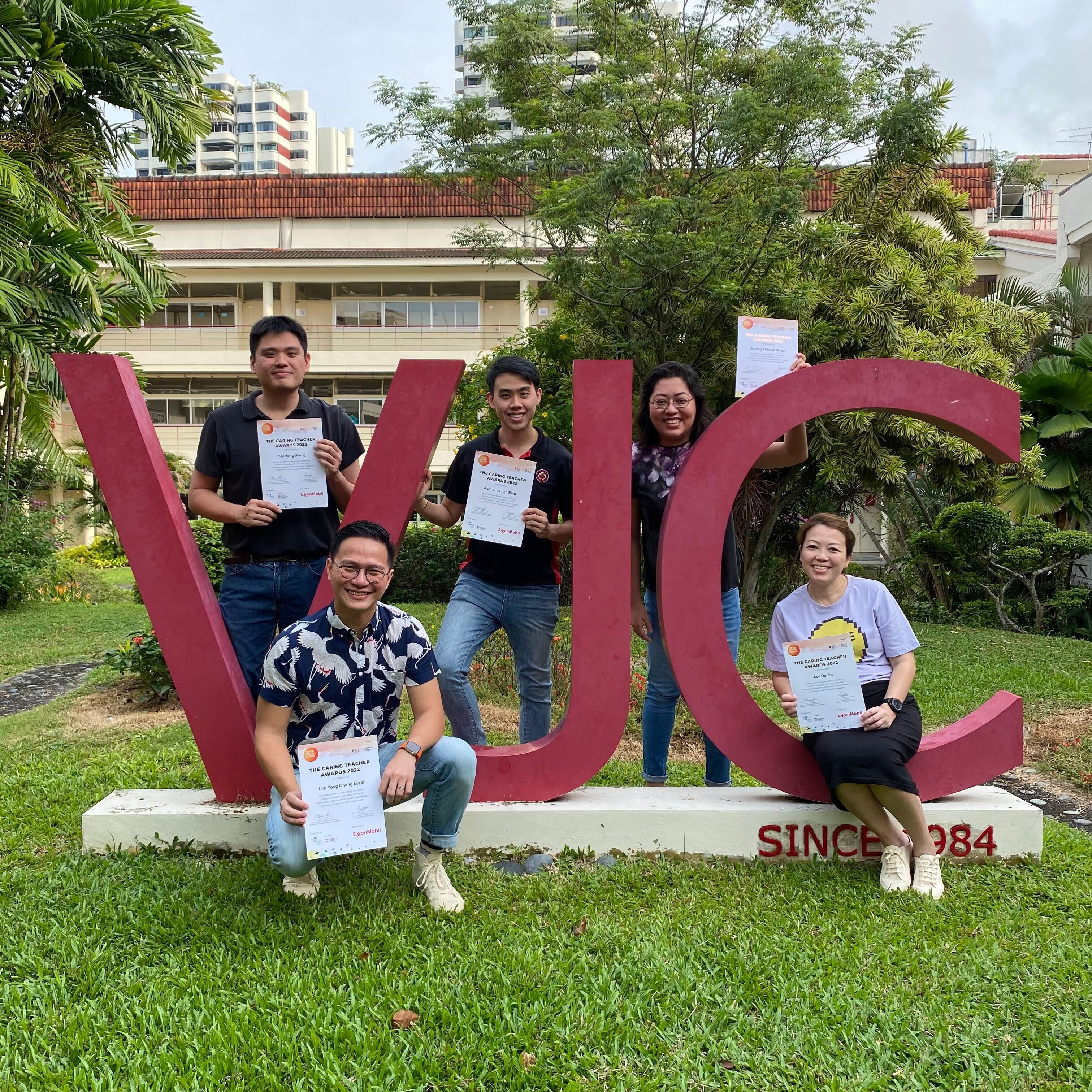 Five people holding certificates pose in front of large red VJC letters on grass, with buildings and trees behind them.